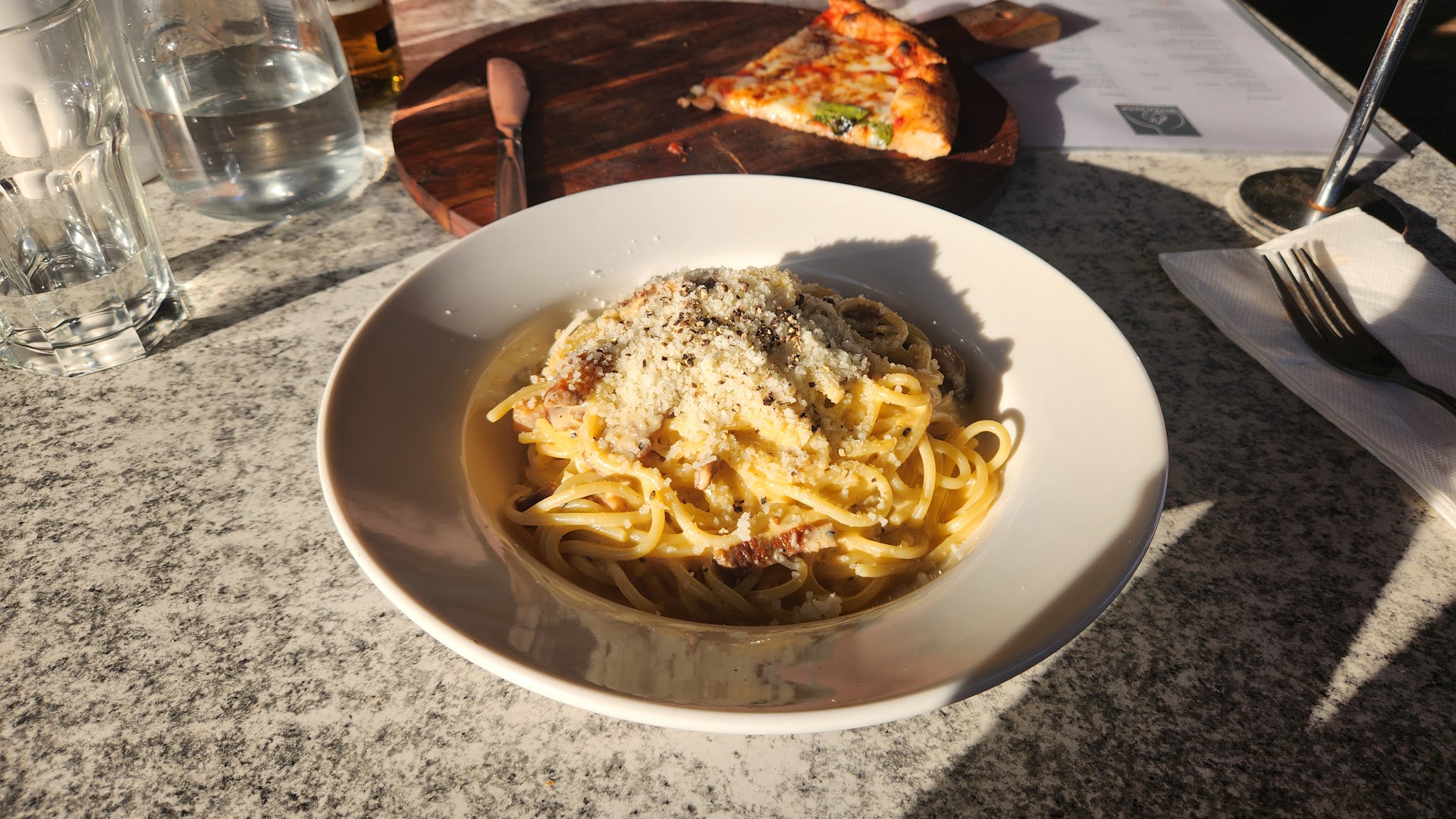 Carbonara and a slice of wood-fired pizza on a sunlit balcony table